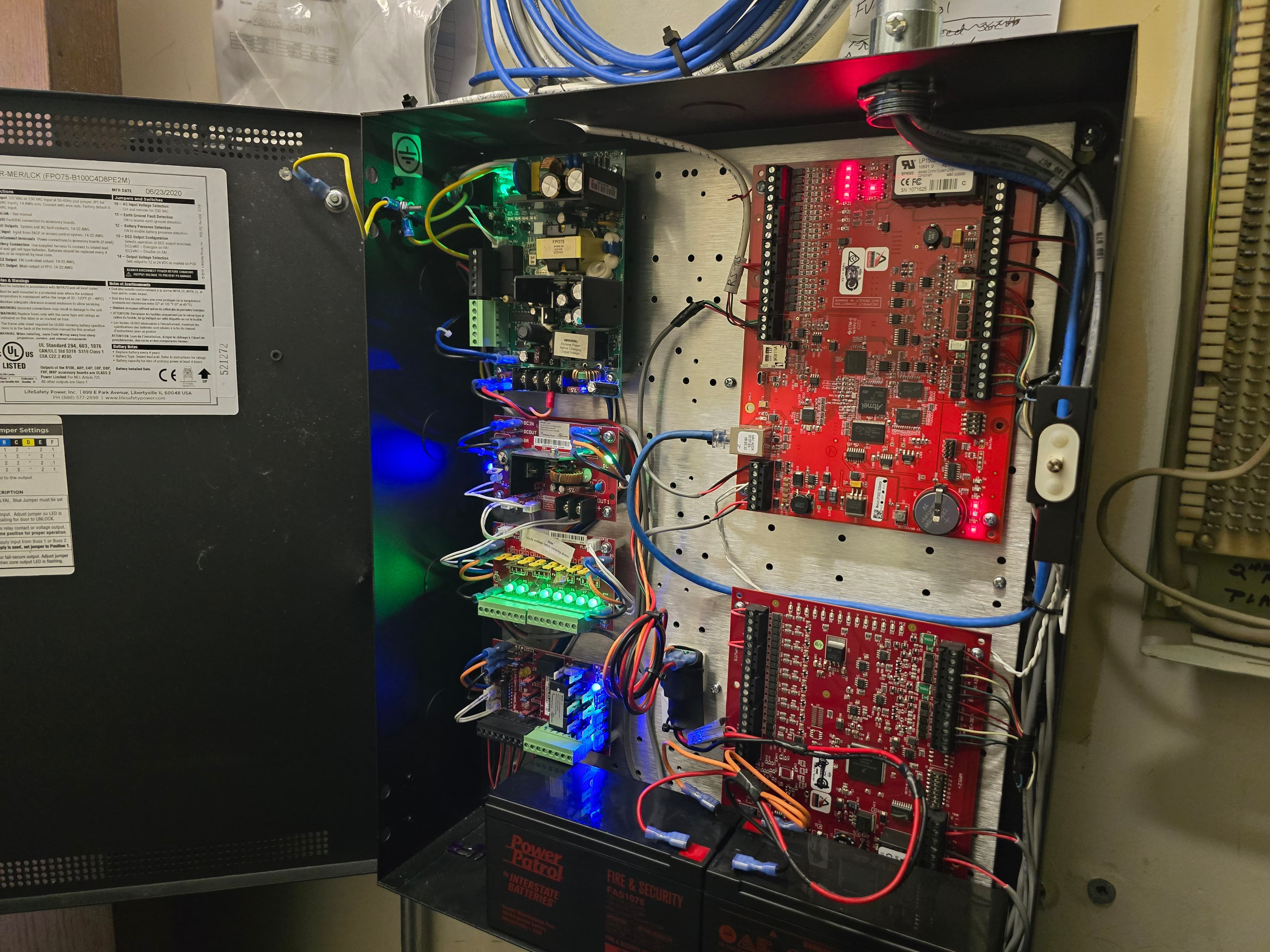 Open metal enclosure with red circuit boards, colorful wiring, glowing LEDs, and backup batteries.