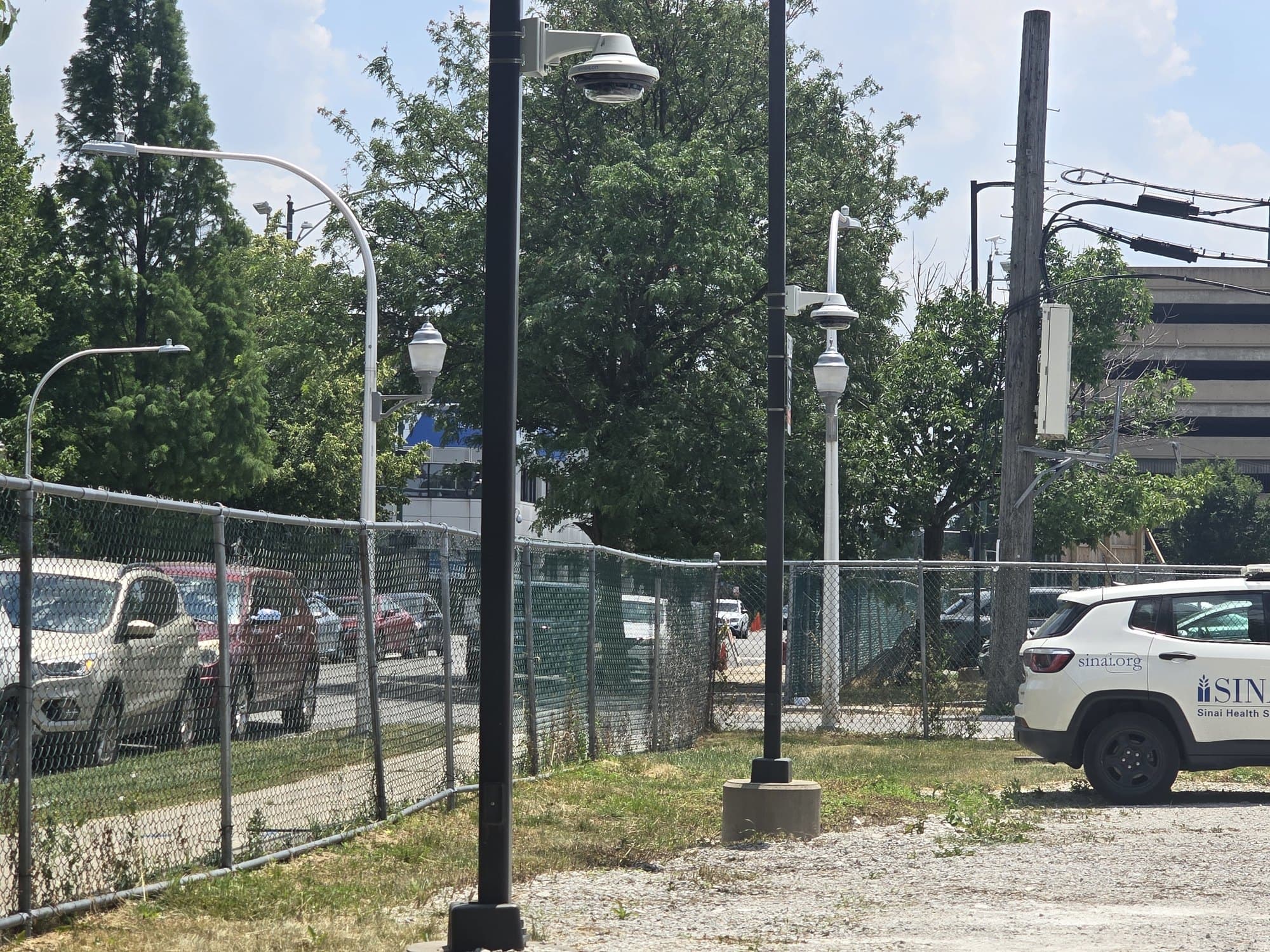 Security cameras on poles overlook a fenced area with a Sinai Health System SUV.