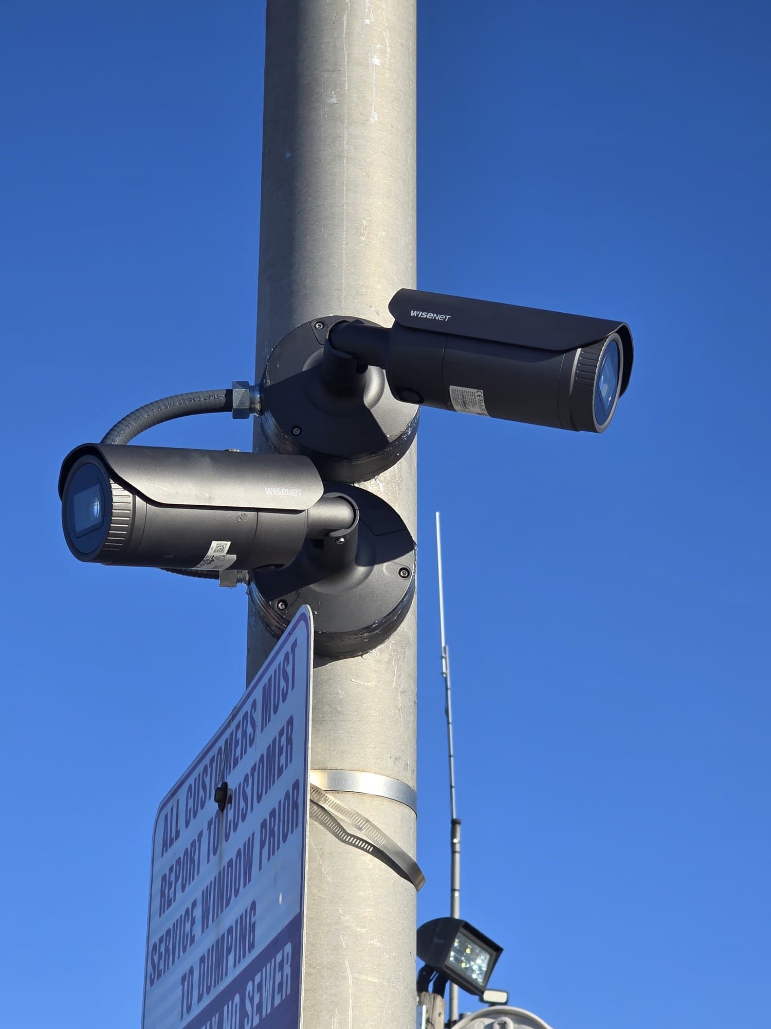 Two black Wisenet security cameras mounted on a grey pole against a bright blue sky.