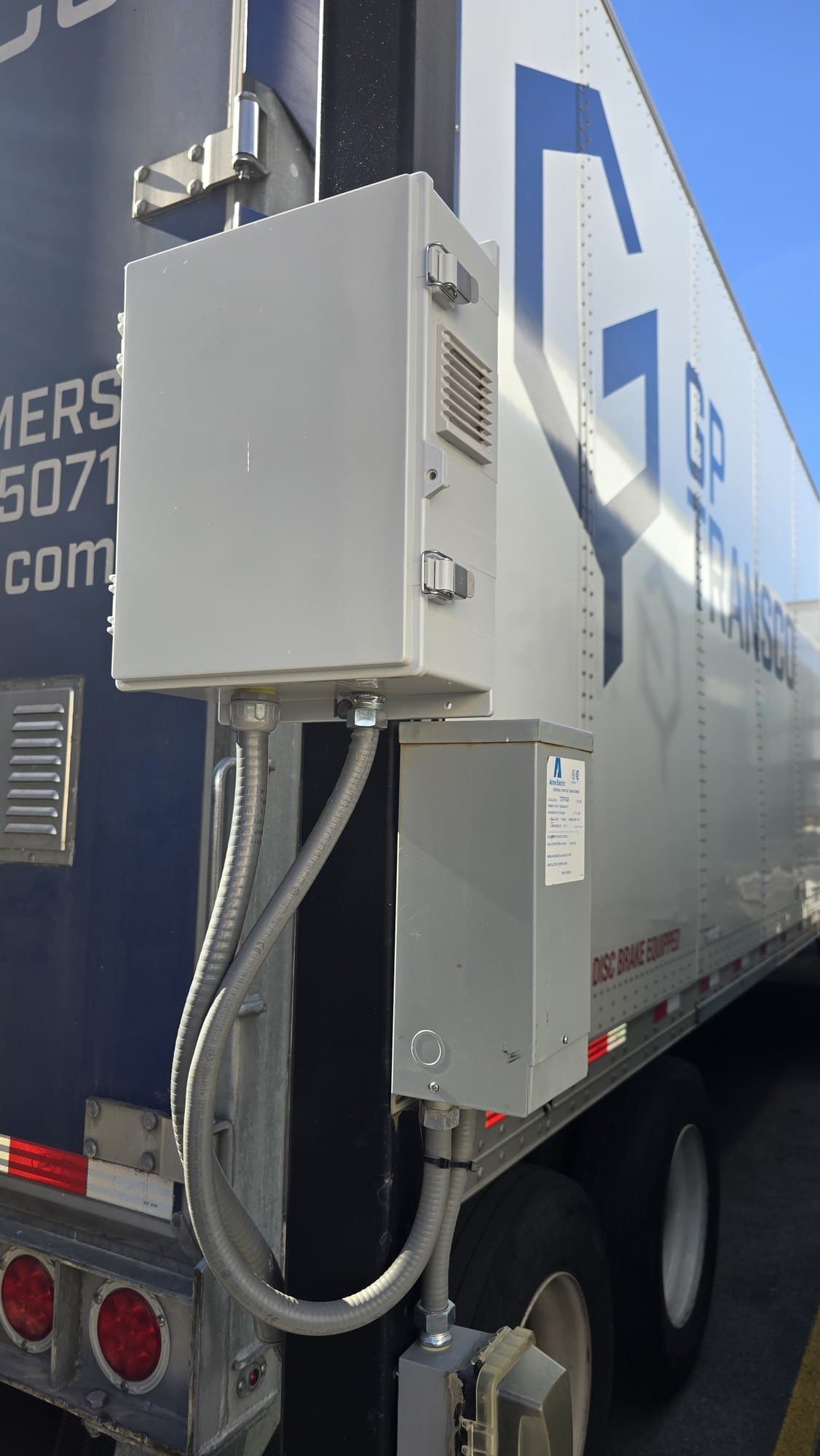 Grey electrical boxes with flexible conduits mounted on the rear of a white semi-trailer.
