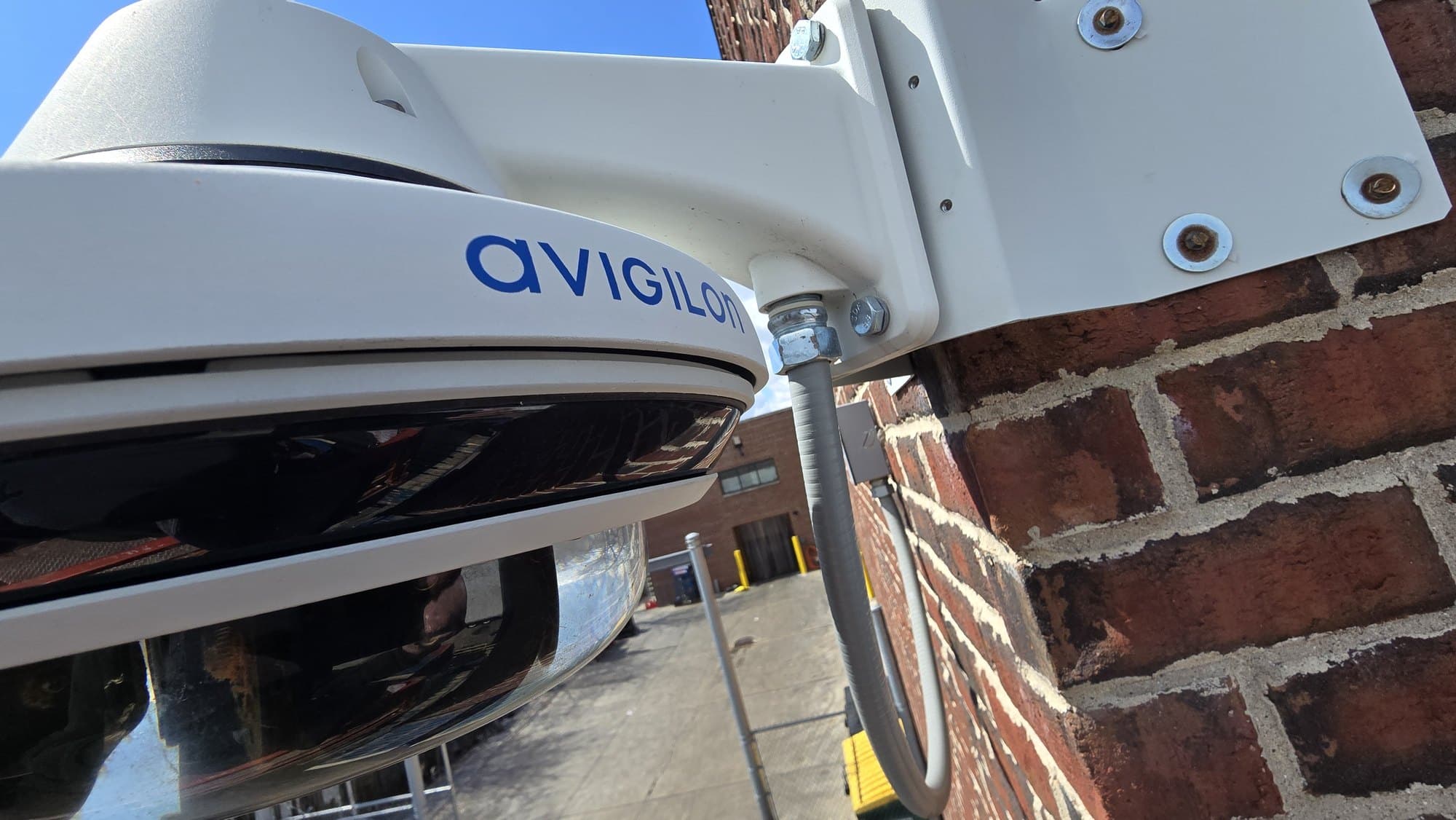 White Avigilon dome security camera mounted on a red brick wall under a blue sky.