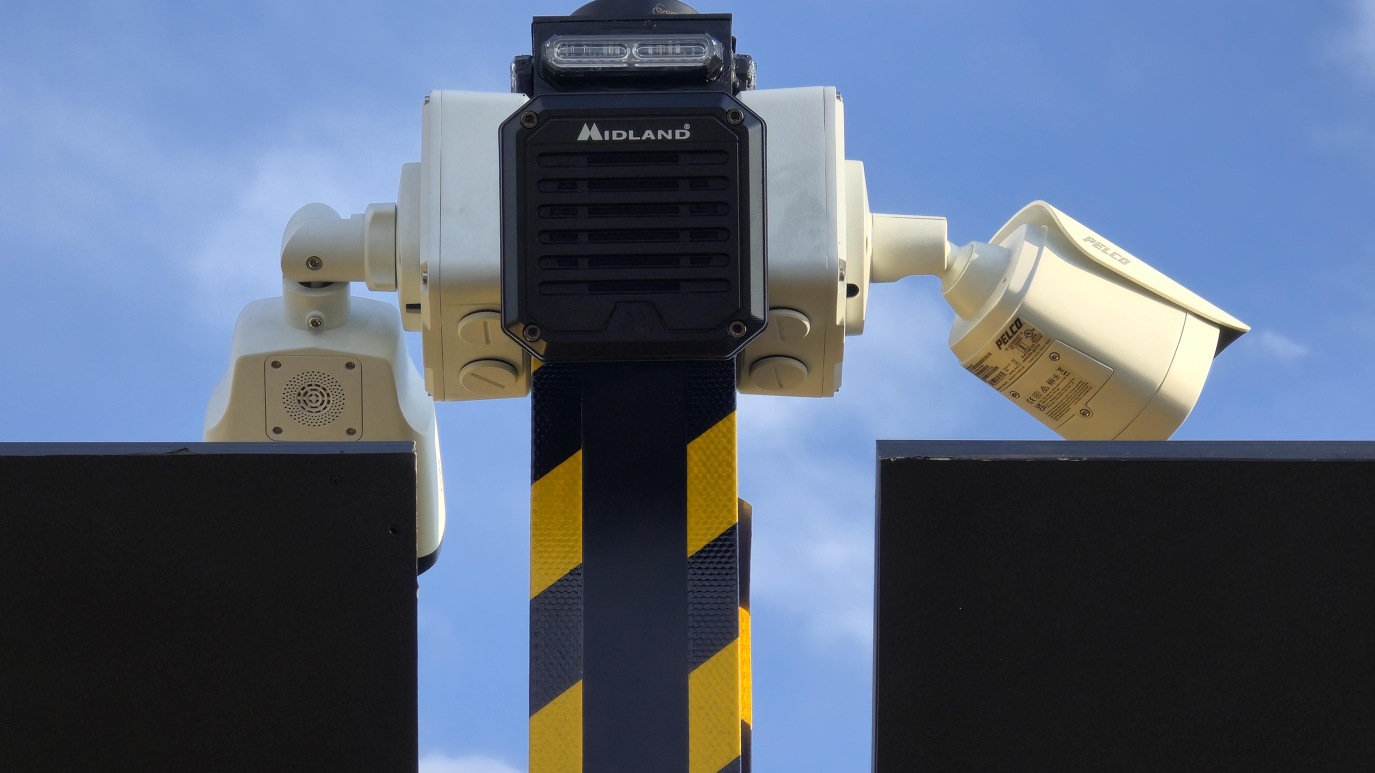 Pelco cameras and Midland speaker on a yellow and black striped pole against blue sky.