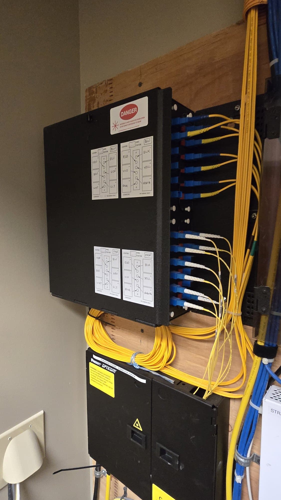 Wall-mounted black fiber optic patch panel with numerous yellow cables and handwritten labels.