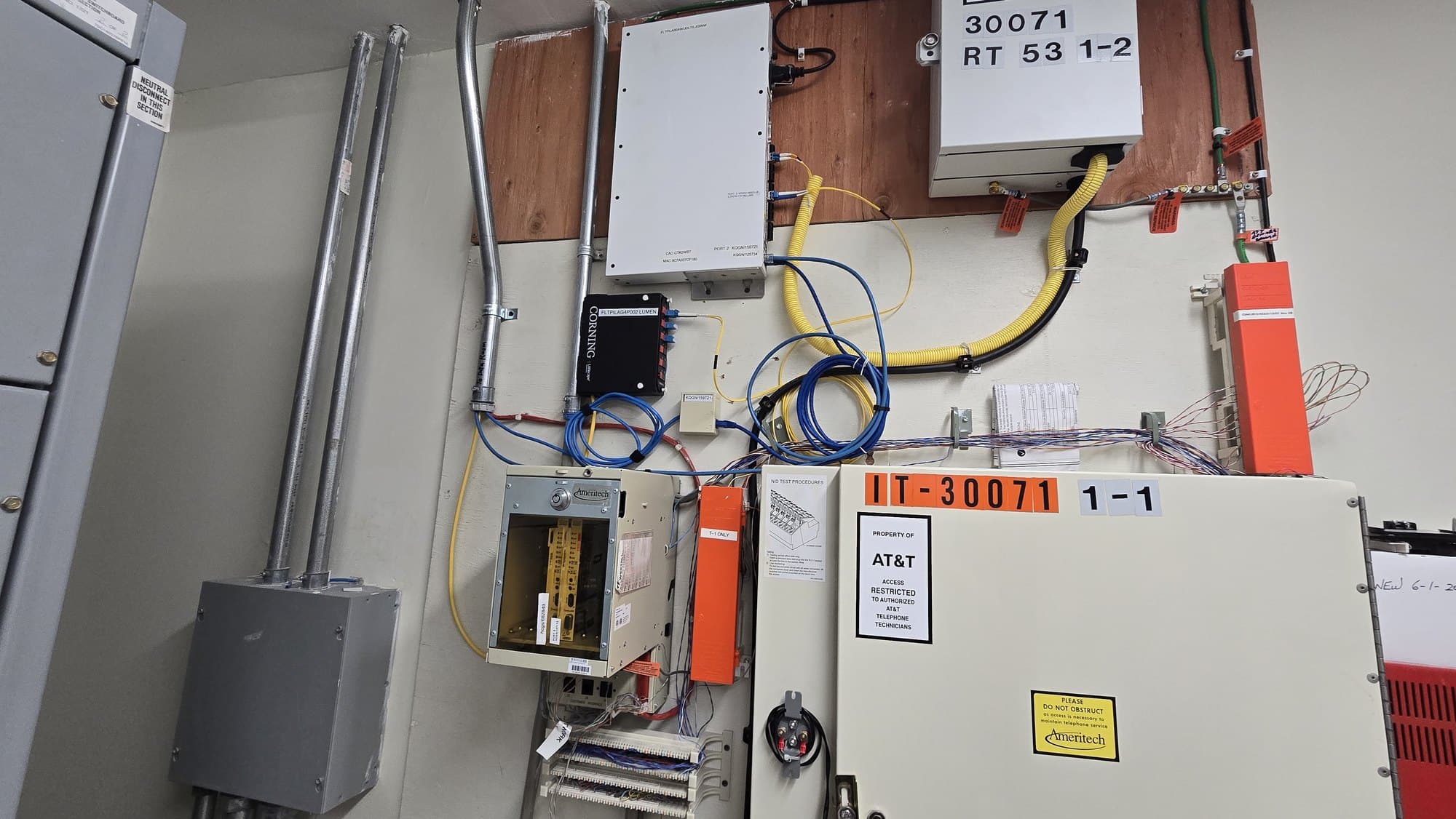 Wall-mounted telecommunications equipment with various boxes, cables, and conduits in an industrial setting.