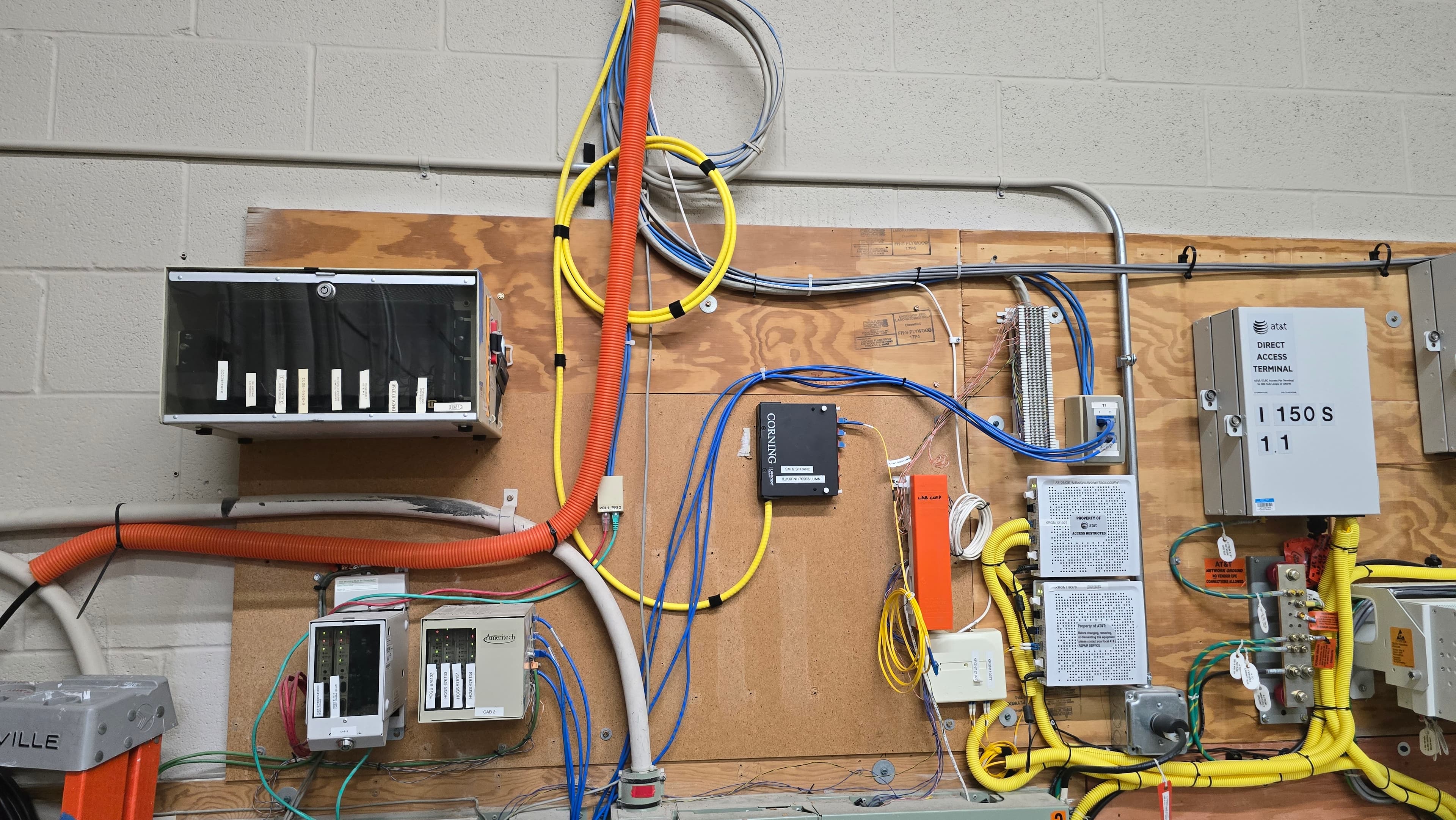 Complex telecommunications equipment and colorful wiring mounted on a plywood board against a white wall.