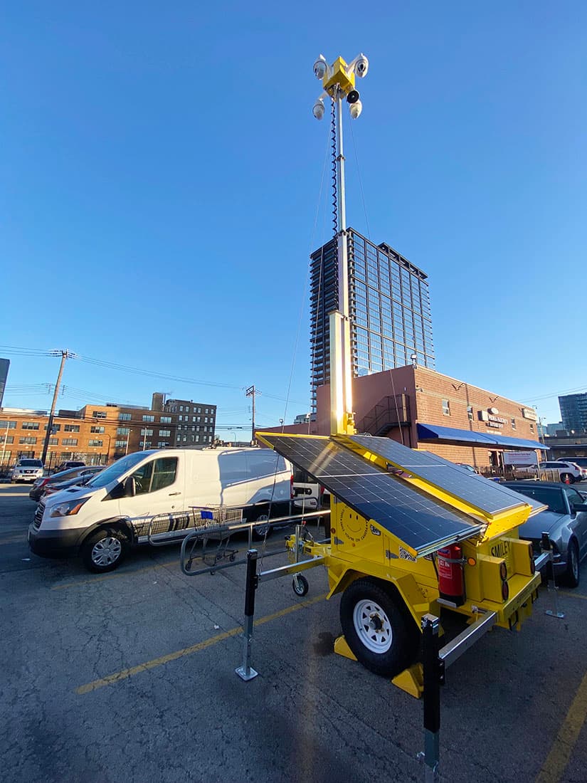 Yellow solar-powered mobile security trailer with a tall mast and cameras in a parking lot.