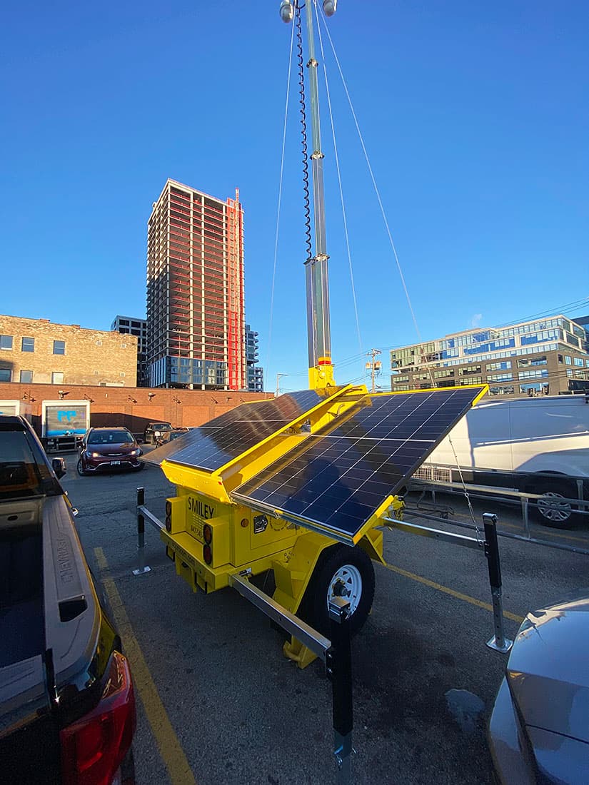Yellow mobile security trailer with solar panels and a tall mast in a city lot.